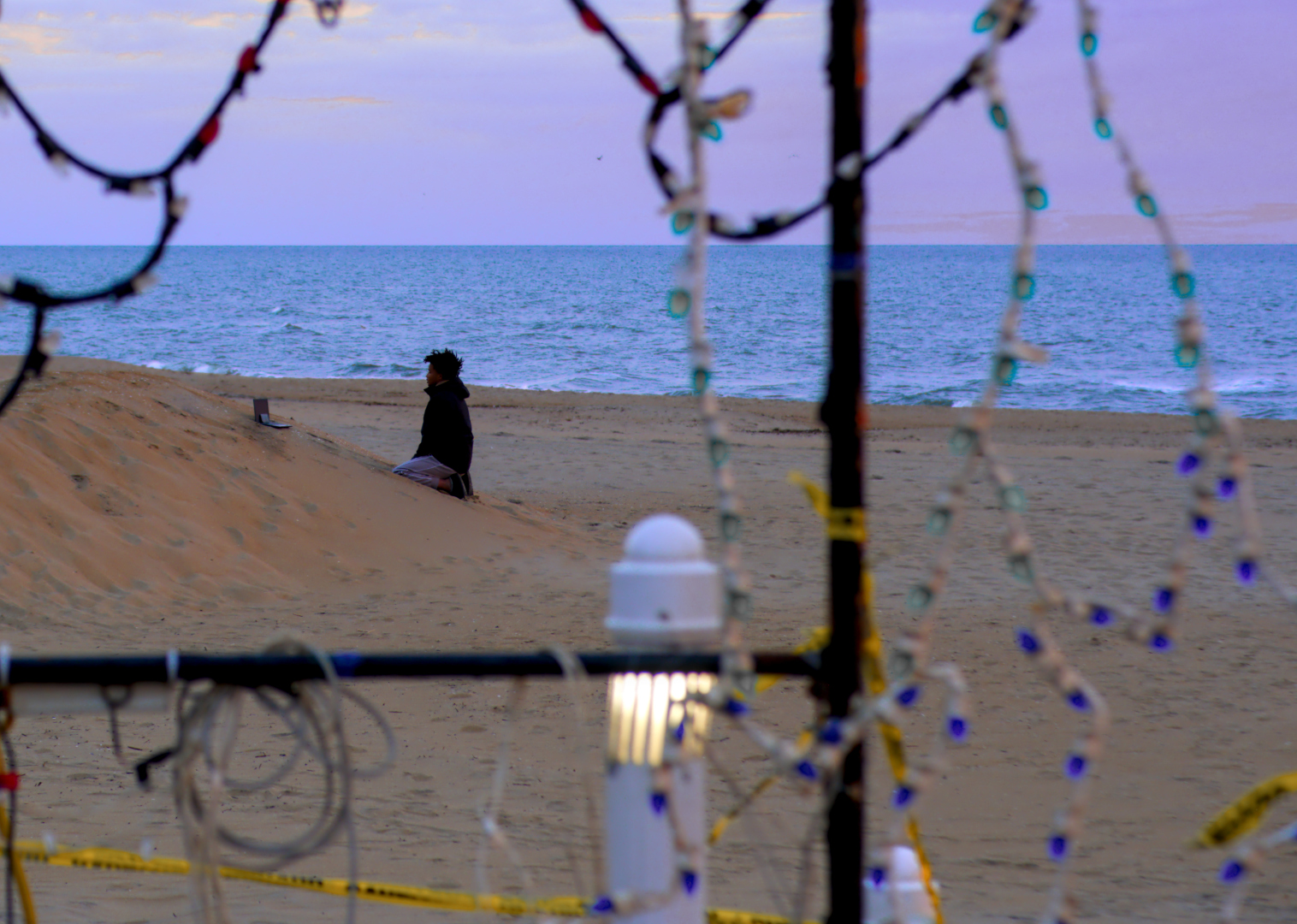 A young man kneeling on a the side of a pile of sand on the beach, apparently looking at his laptop and phone which are sitting in front of him on the slope.