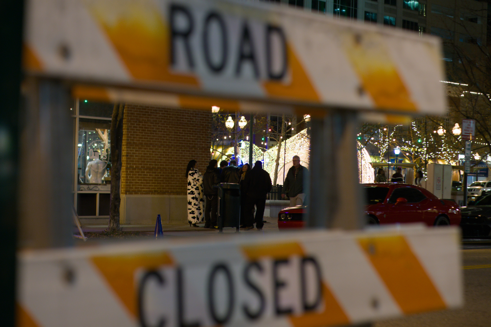 A group of people wrapped in warm winter clothing, waiting for entry to a restaurant or club in the evening. There are Christmas light display nearby. We see the group through the horizontal parts of a road closure sign with bright orange markings. The scene is punctuated by the woman wearing a gorgeous black and white faux fur jacket in a sea of otherwise dark clothing.