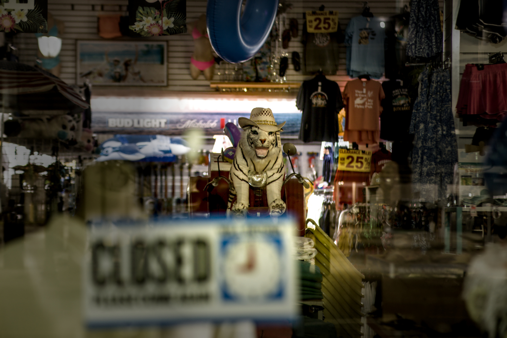 Early morning shot of a store selling an eclectic variety of goods. Highlighted at the center of the shot is a large stuffed white tiger with black stripes and a ha, seated upon a toy motorbike. The tiger is surrounded by items like an inner-tube, beachwear, advertisements for beer, and many other items. Barely readable is a "Closed" sign on the door showing that the proprietor will return by 9 a.m.